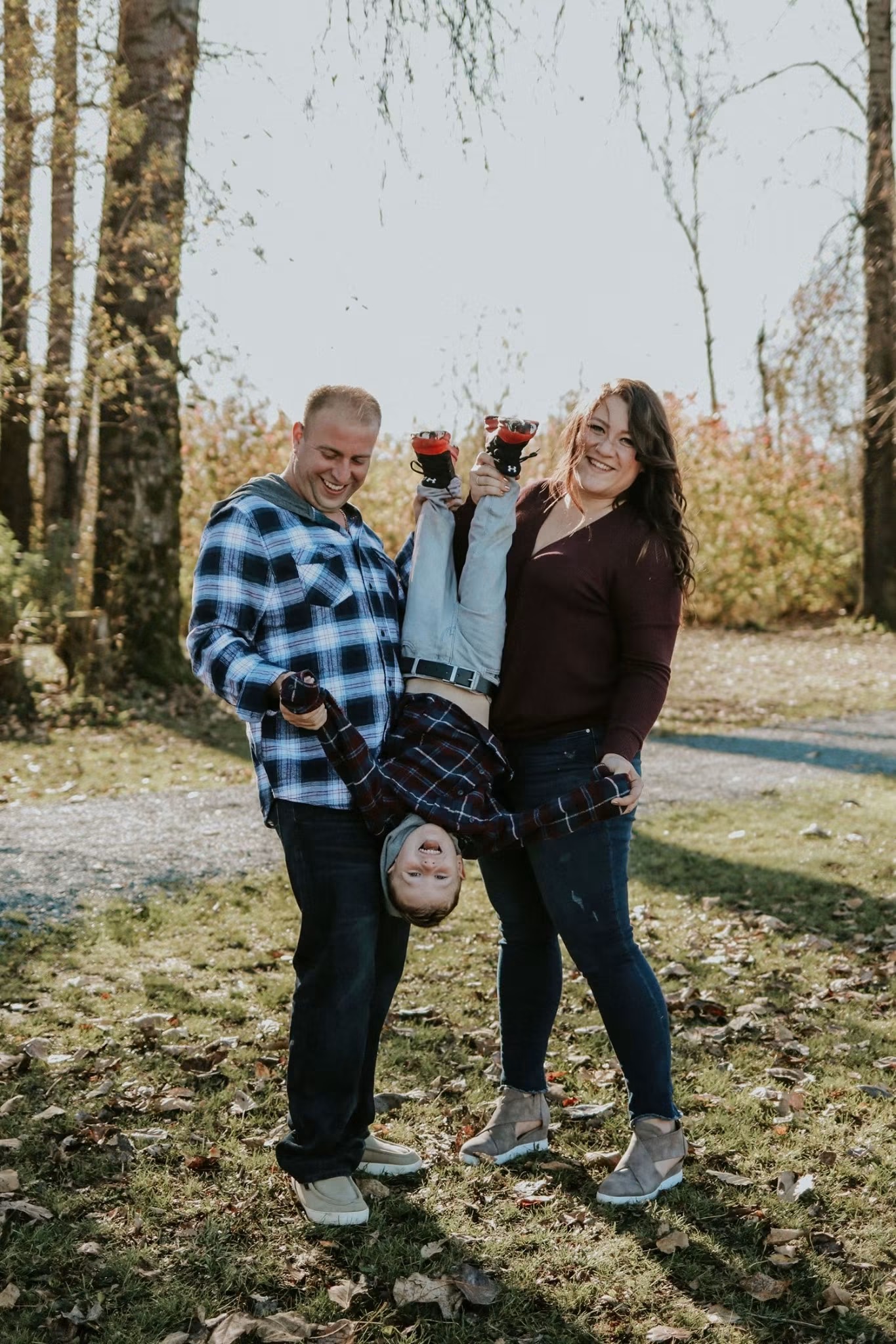 Parents laughing as they swing their young son upside down in autumn light