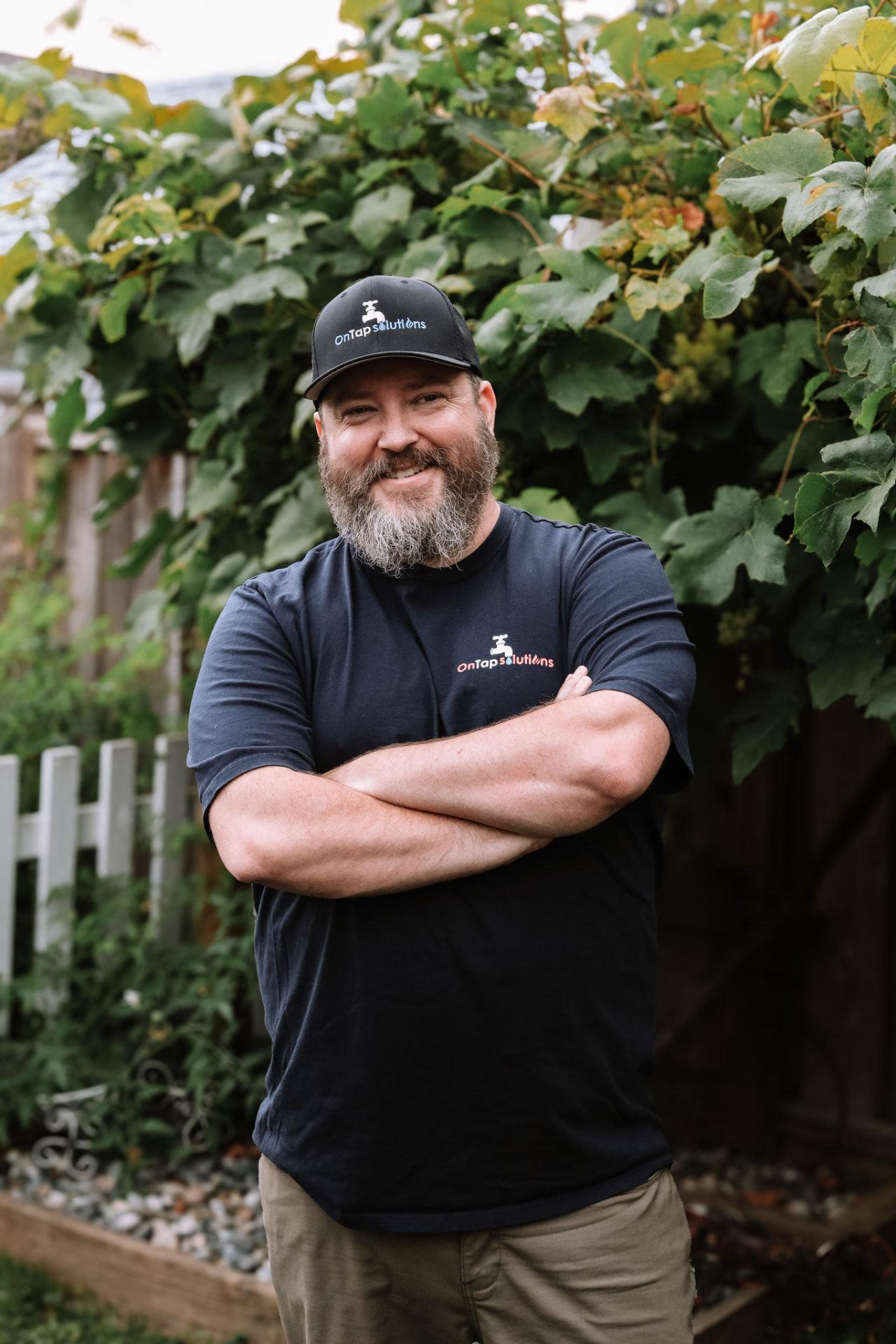 A smiling bearded man in a navy shirt and cap standing with arms crossed in a garden