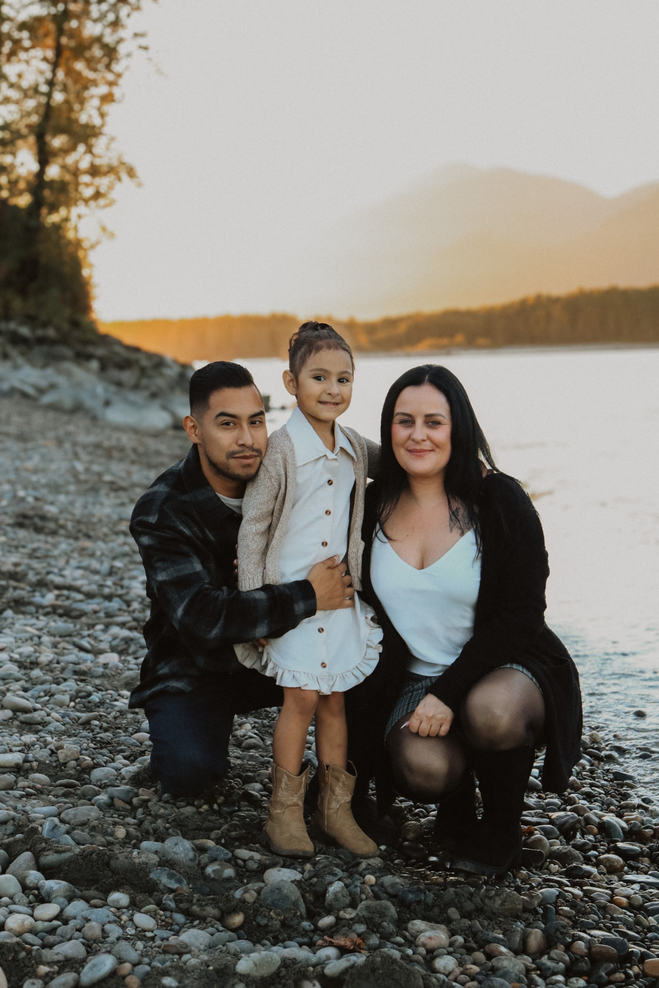 Family of three crouched together on a pebble shore at sunset
