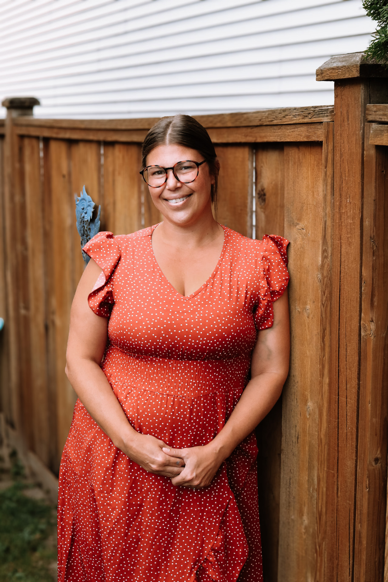 A smiling woman in an orange polka-dot dress leaning against a wooden fence