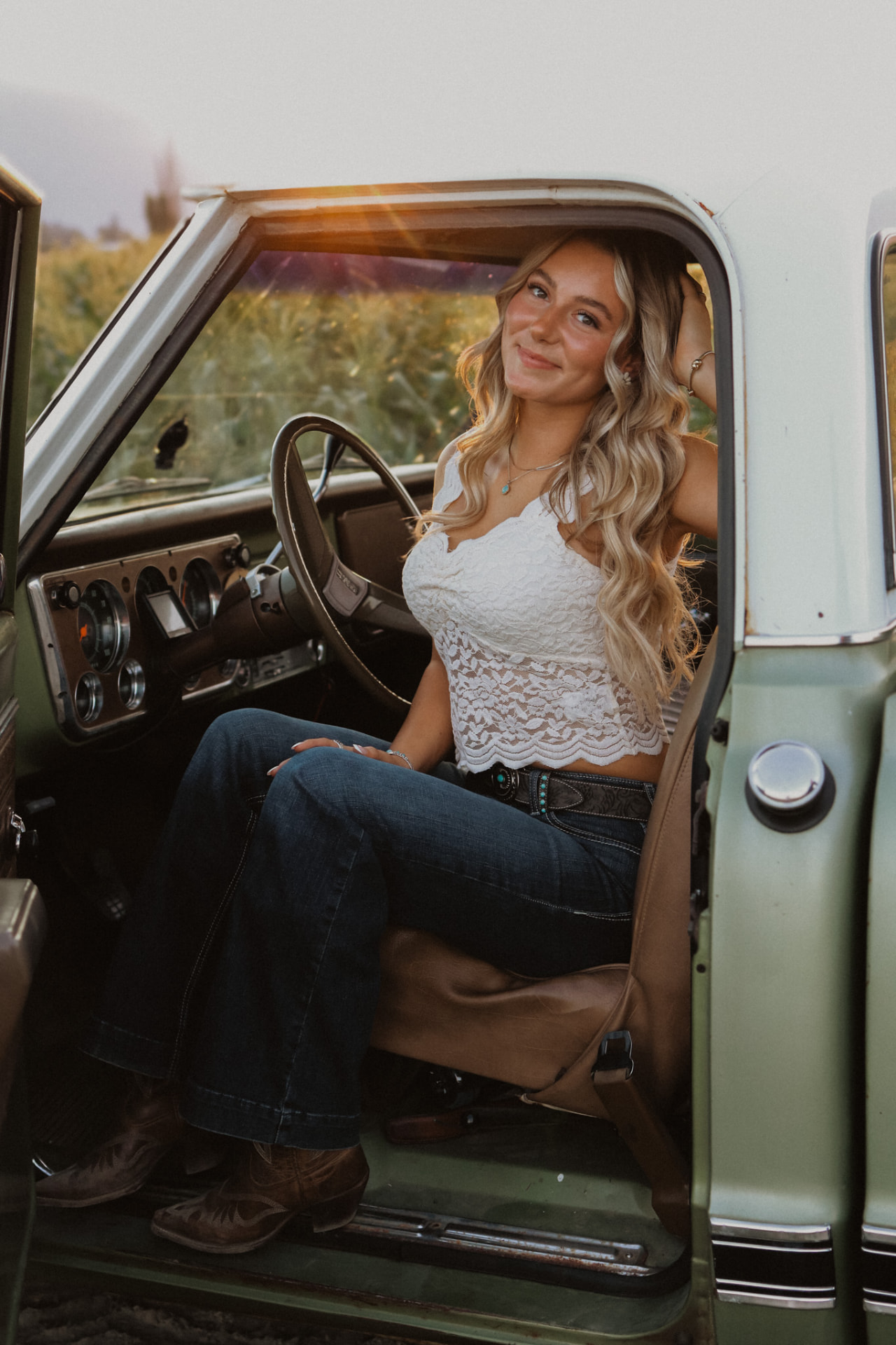 A woman in a white lace top and jeans sitting in the cab of a vintage green GMC truck at sunset