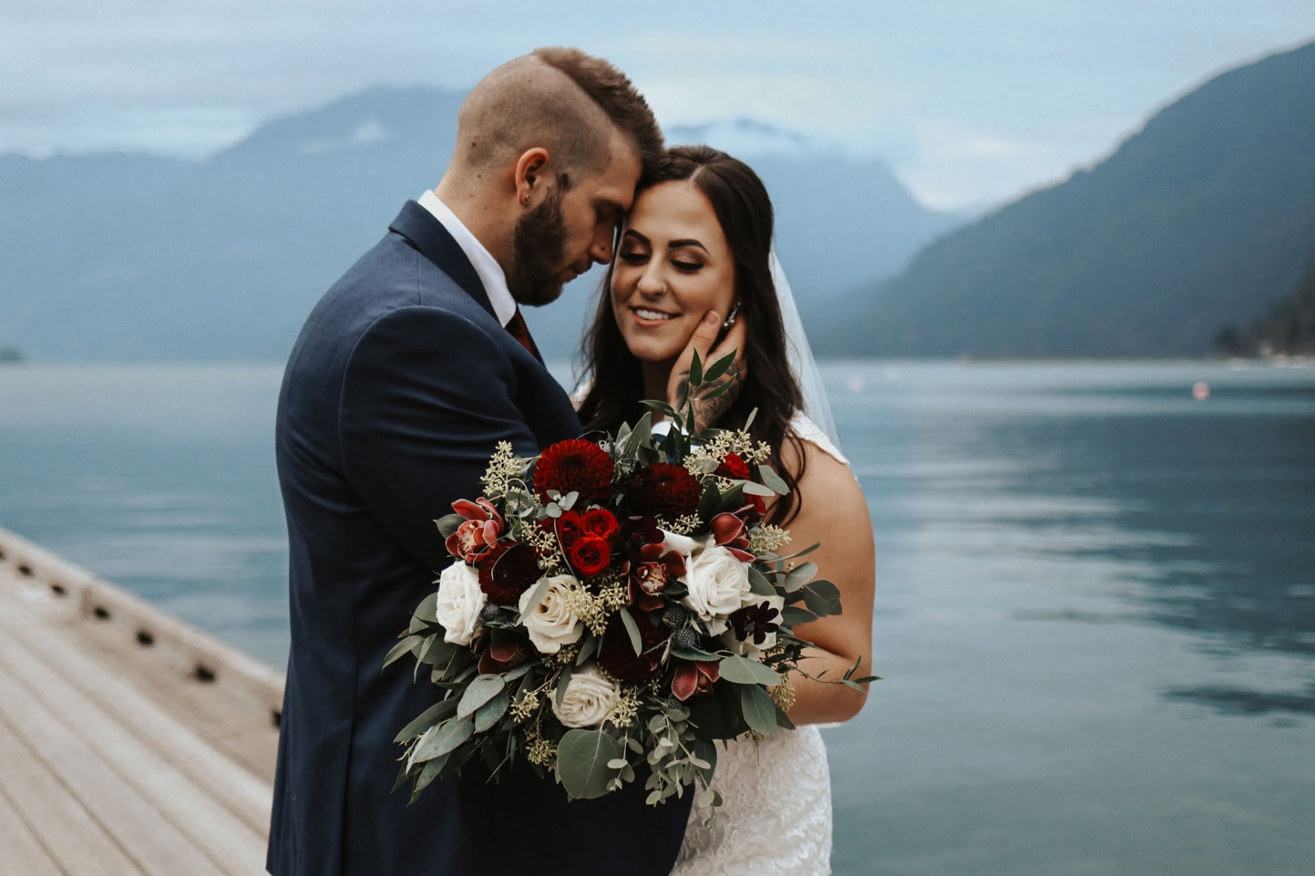 A bride and groom foreheads together on a lakeside dock with a deep red bouquet