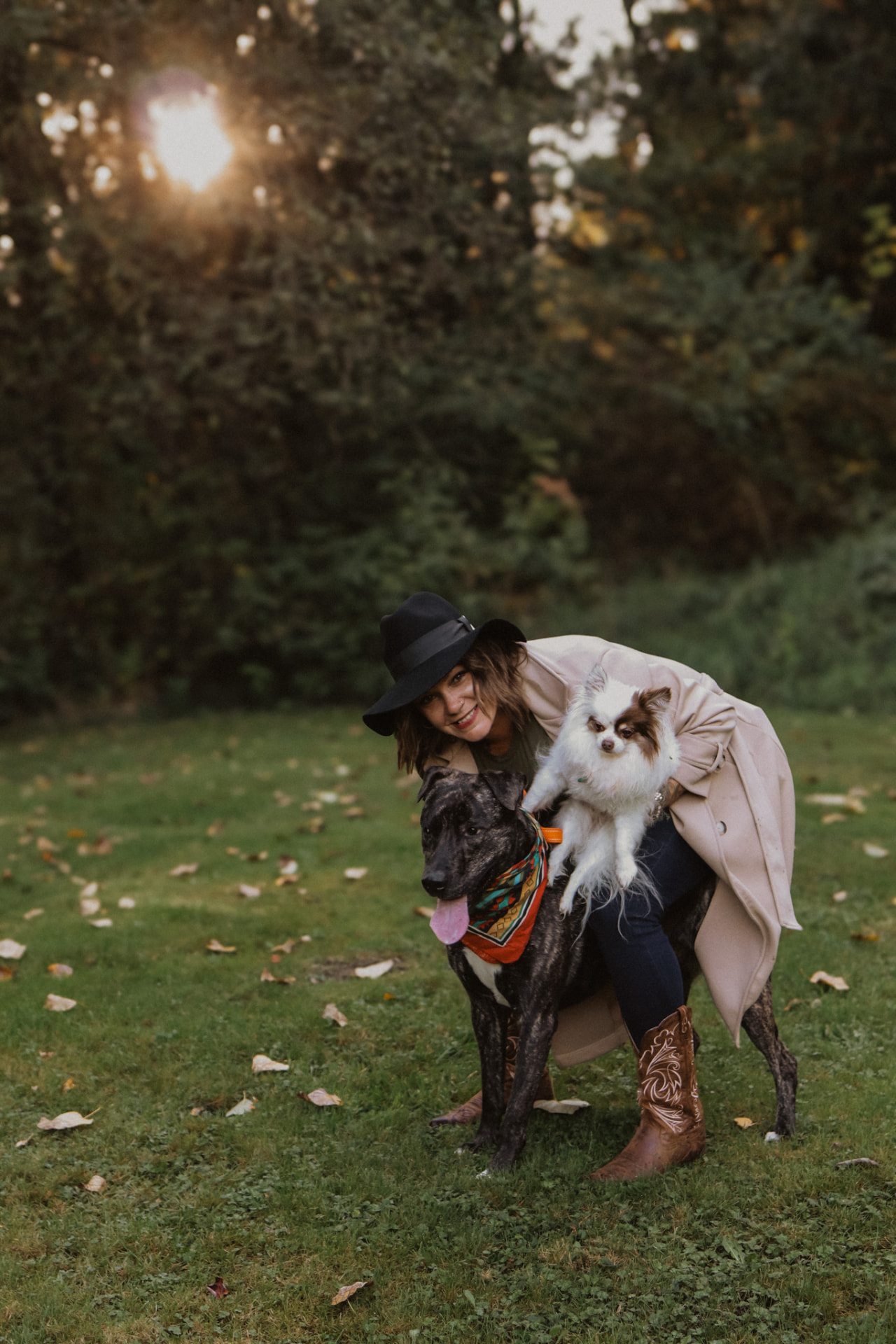 Woman with a black hat hugging a brindle dog and holding a Pomeranian at golden hour